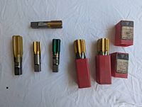 Photo showing six machinist taps of different coatings and sizes, with two in red plastic cases and two cardboard boxes containing taps.