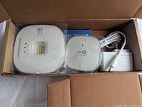 Overhead view of the Chamberlain MyQ smart garage control kit inside the box. Shows the main control unit, the wireless sensor, power adapter, and manual.