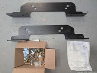 Two black metal mounting brackets placed flat on surface with hardware box below them containing bolts, nuts, washers.