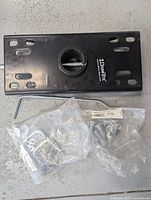 Black rectangular hitch head with central circular ball mount and Draw Tight branding, accompanied by metal rod and accessories in plastic bags, placed on concrete floor.