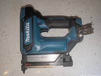 Side view of Makita PT354D cordless pin nailer lying on white countertop, showing blue and black coloring and Makita logo
