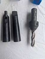 Photo showing the two black metal Morse taper adaptors and the bit holder with drill bit attached lying on white cloth surface.