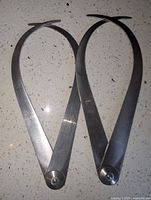 Two Starrett outside calipers side by side showing their curved leg shape, pivot points, and adjusted positions on a light speckled surface.