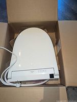 Top-down view of the white Toto SW3083 bidet seat in its cardboard packaging, showing the seat lid and attached water hose.