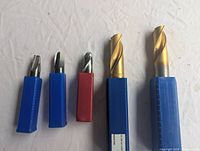 Photo showing five metal milling cutters with cylindrical shanks in blue and red plastic cases, two cutters have gold coated cutting edges.