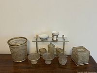 Full set of gold-toned bathroom accessories on tabletop including tissue box cover, soap dispenser, soap dishes, small decorative bowls, and hanging shelf.