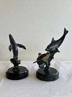 Two bronze sculptures — whale and dolphin — shown on flat surface against a plain wall, capturing the full view of both sculptures and their marble bases.