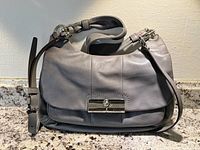 Front angle view showing soft grey leather, silver-tone clasp with Coach branding, flap closure, and adjustable straps.