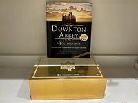 Photo showing the companion book titled 'Downton Abbey A Celebration' placed behind the golden collector's box featuring an illustration of Downton Abbey.