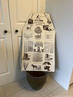 Front view of antique style gardening apron displayed on pottery planter, showing the detailed black print gardening illustrations on cream fabric apron, with waist and neck ties.