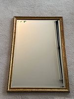 Full view of the rectangular wall mirror with gold frame and beveled glass against a carpeted floor.