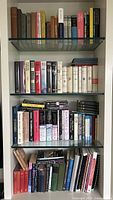 Four shelves of assorted hardcover and paperback books including a mix of genres and authors, showing spines and some covers.