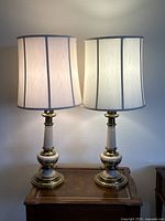 Pair of vintage Stiffel Hollywood Regency style table lamps with brass and white enamel bases, topped with white fabric drum shades, shown side-by-side on wooden tables.