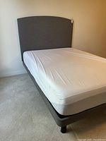 Front perspective of grey upholstered double bed frame with white mattress cover, showing curved headboard and platform base on four round black legs.