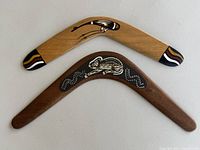 Pair of two wooden boomerangs displayed flat. Smaller light wood boomerang with painted tribal designs and larger darker boomerang with lizard dot painting.
