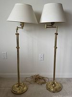 Pair of two mid-century brass swing arm floor lamps shown side by side. Lamps have white fabric shades and visible patina on brass.