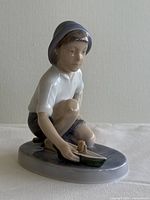 Side angle view of the Royal Copenhagen ceramic figurine showing the boy squatting while holding the toy boat on an oval base.