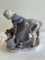 Front-left angle image of the porcelain figurine showing the woman bending and feeding cow from bucket.