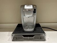 Front view of the silver Keurig coffee maker with clear water reservoir and wire frame storage drawer below.