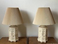 Pair of cream floral porcelain lamps with beige pleated lampshades on wooden surface against a white wall.