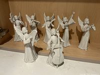 Photo of all 7 white Dresden porcelain musical angel figurines displayed on shelf, showing each angel holding a different instrument.