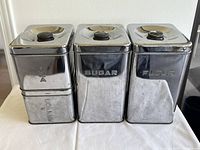 Front view of four Lincoln Beauty Ware chrome canisters labeled Tea, Coffee, Sugar and Flour, showing chrome finish and shapes.