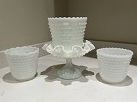 Four white vintage milk glass pieces on a countertop: two small hobnail planters, one pedestal dish with ruffled edge, and one smaller hobnail container