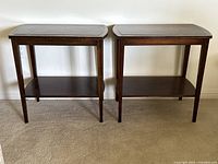 Pair of vintage wooden side tables with leather inlaid tops and lower shelves placed side by side on carpeted floor.