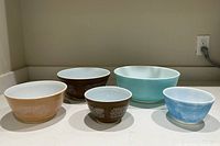 Five vintage Pyrex mixing bowls, three Brown Woodland brown with white floral patterns and two blue bowls with white interiors, arranged on a countertop.