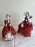 Pair of vintage Royal Doulton female figurines in red dresses and bonnets, one holding a white lantern accessory.