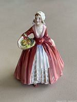 Front view of Royal Doulton Janet figurine showing red dress, white apron and basket of yellow flowers.