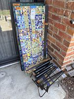 Stacked three mosaic tabletops leaning against brick wall with removed legs lying nearby