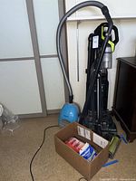 Photo showing light blue Panasonic canister vacuum with attached gray hose and wand held upright against wall. A box with vacuum bags and accessories is on the floor nearby.