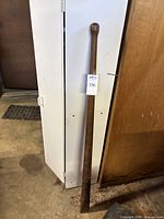 Full length view of a 53-inch tall wooden handle leaning against a cabinet, showing wedge-shaped end and rounded knob end.