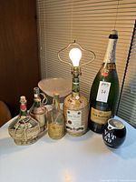 Photo of Bacardi lamp with bulb lit, multiple decorative bottles including a champagne bottle, a wicker wrapped bottle, and a small black spherical bottle.