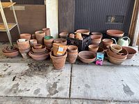 Wide view of multiple stacks and loose piles of mainly clay pots, with some plastic pots and a watering can. Items show dirt and outdoor use with some discoloring.