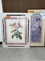 Two framed art prints side by side on floor against a door: left is pink rose botanical print with pink marble border, right is lavender Alphonse Mucha print.