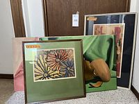 Lot of several art pieces stacked against a wall including framed floral print, figurative print, and unframed still life painting.