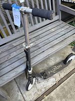 Fuzion Cityglide kick scooter leaning against a wooden bench. Visible scuffs and minor scratches on the frame and deck. The scooter has white wheels and black deck with Fuzion branding.