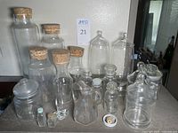 Multiple clear glass vessels ranging from small bottles to large jars with cork and glass lids arranged on a surface against a wall.