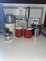 Photo showing entire lot including Krups coffee grinder, Bialetti espresso maker boxes, handheld frother, red coffee pots, glass jug, stainless steel pitcher, and metal strainer.