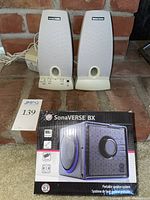 Pair of Harman Kardon SV Solo speakers shown from front, with fabric grilles and Harman Kardon branding clearly visible. Control knobs for bass, treble, and volume visible on the left speaker.