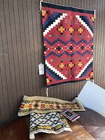 Full view of hanging woven textile on wall and two needlework pieces on table below.