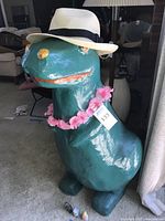 Large green paper mache dinosaur standing upright wearing white hat and pink lei