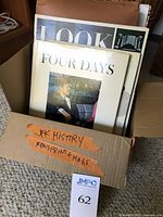 Box labeled 'JFK History, Newspaper + Mags', containing several magazines and newspapers visible from top, including Kennedy related material, with orange tape labeling