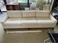 Front view of three-seat light fabric sofa showing thick cushions and visible stains on upholstery. Sofa sits on wooden legs with a base frame.