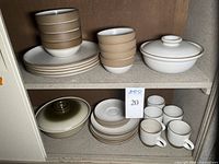 View of two shelves containing stacks of earth-toned ceramic dinnerware including bowls, plates, mugs and lidded dishes.