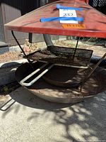 Front side view of the fireplace showing red funnel-shaped enameled tops with item stickers, grill rack with handles, and round fire bowl below, with visible rust and dirt on surfaces.