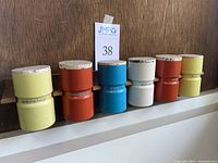 Six aluminum spice jars with colorful paint and cork lids lined up on a white surface in front of a wooden wall, showing various spice labels.