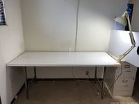 Front view of white laminate top table with chrome legs and adjustable feet, shown with task lamp and partial file cabinet.